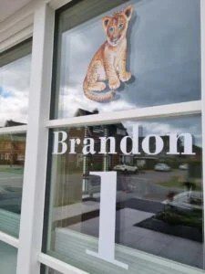 Raamsticker met geschilderde leeuwenwelp met blauwe ogen en witte tekst Brandon op etalageruit