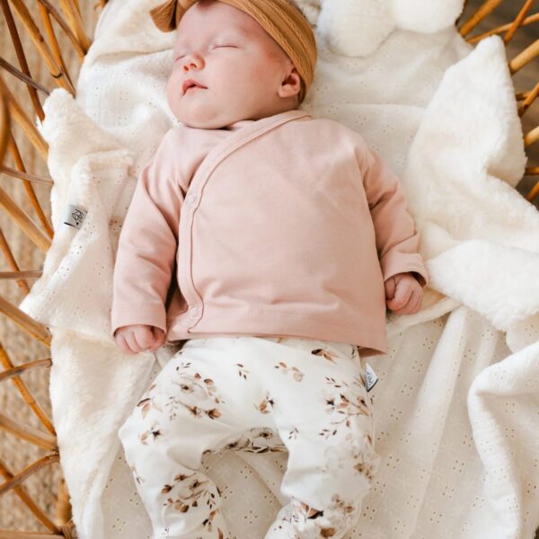 Slapende baby in rotan wiegje met roze shirt, gebloemde broek en strik haarband naast pluche schapenkussentje