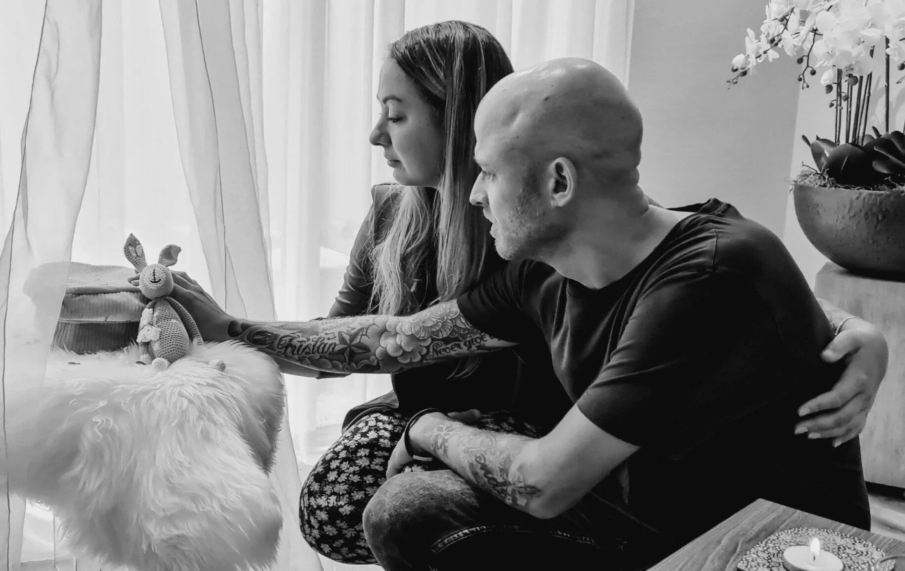 Man en vrouw met getatoeëerde armen bekijken een baby in gezellige woonkamer met witte gordijnen en orchideeën
