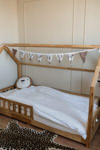 Houten huisjesbed met wit gestippeld dekbed, naamslinger en knuffel in neutrale babykamer