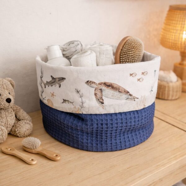Opbergmand met zeeleven-print gevuld met handdoeken en babyborstels op houten commode in babykamer